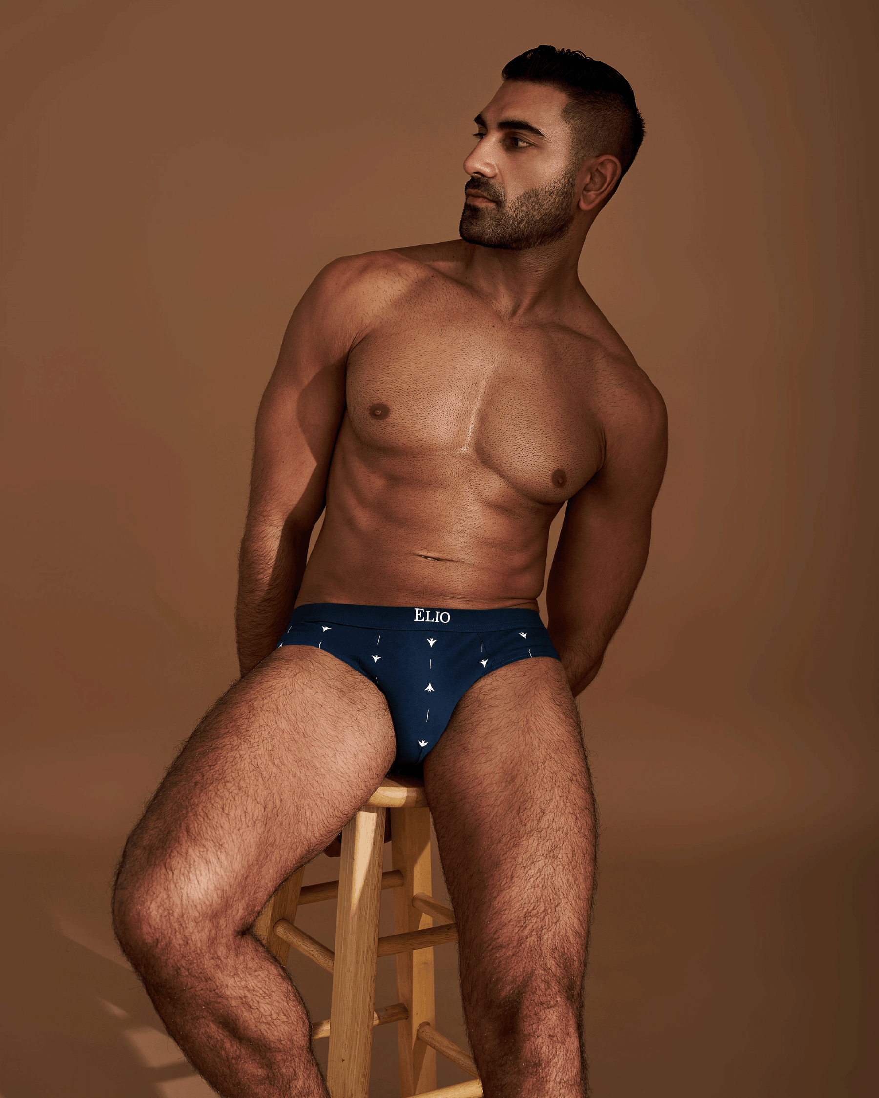 A shirtless man sitting on a wooden stool wearing navy blue briefs with a small white geometric pattern and the brand name 'ELIO' on the waistband, set against a warm brown studio background.

