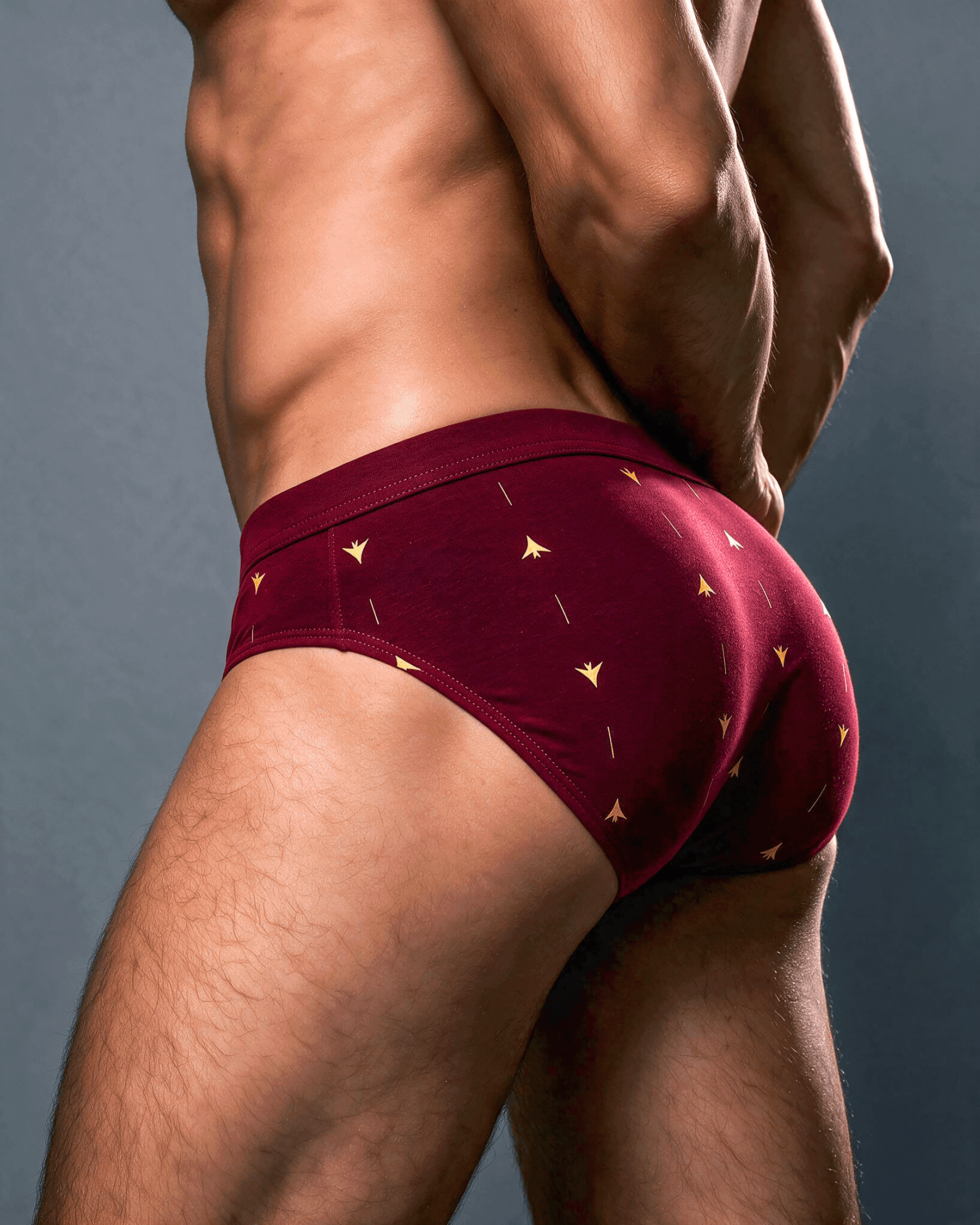 Rear view of a male model wearing burgundy briefs with a small gold geometric pattern, set against a dark gray studio background.