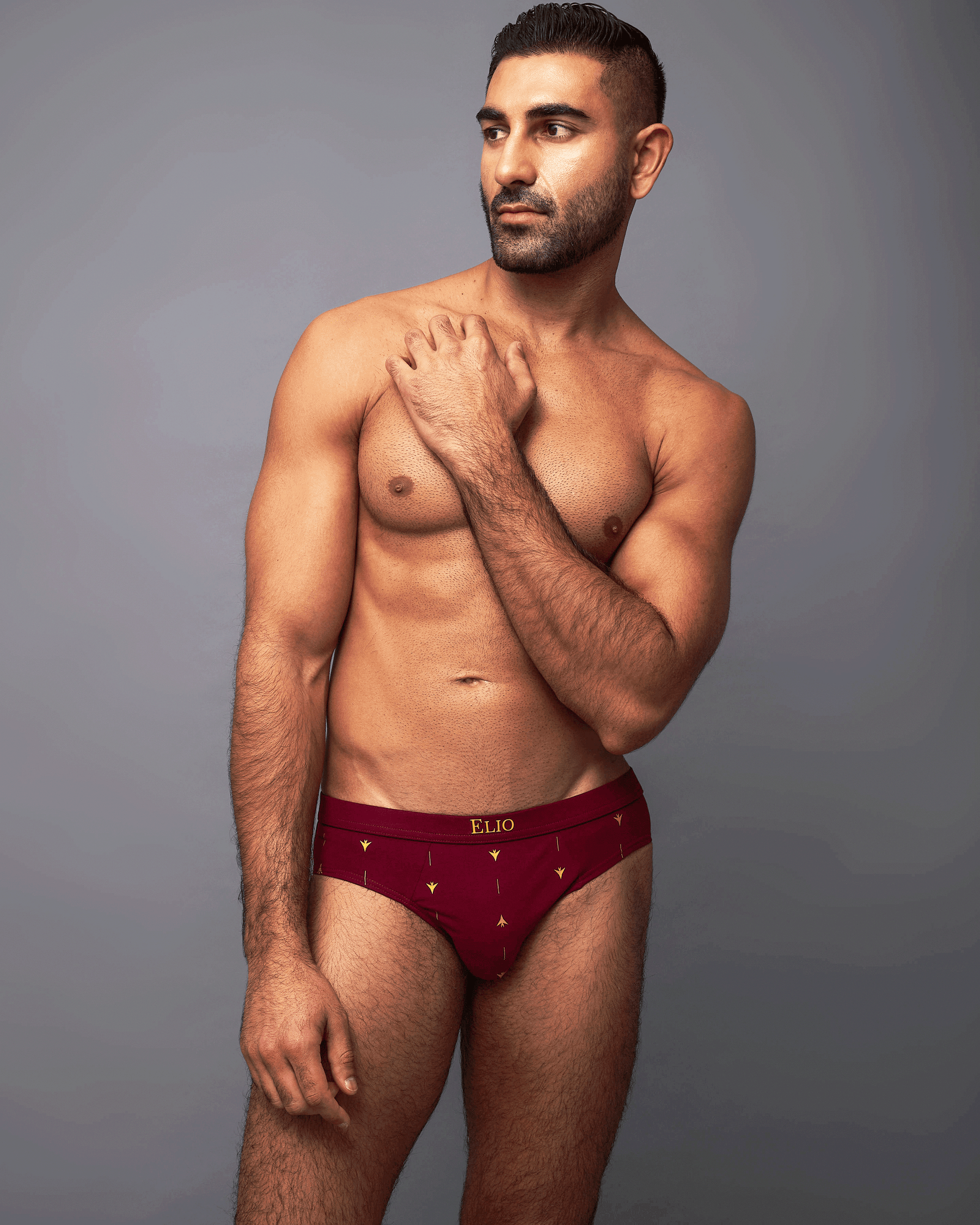 A male model wearing a pair of burgundy briefs with small gold star patterns and a gold ELBO brand label on the waistband, posed against a solid gray studio background.