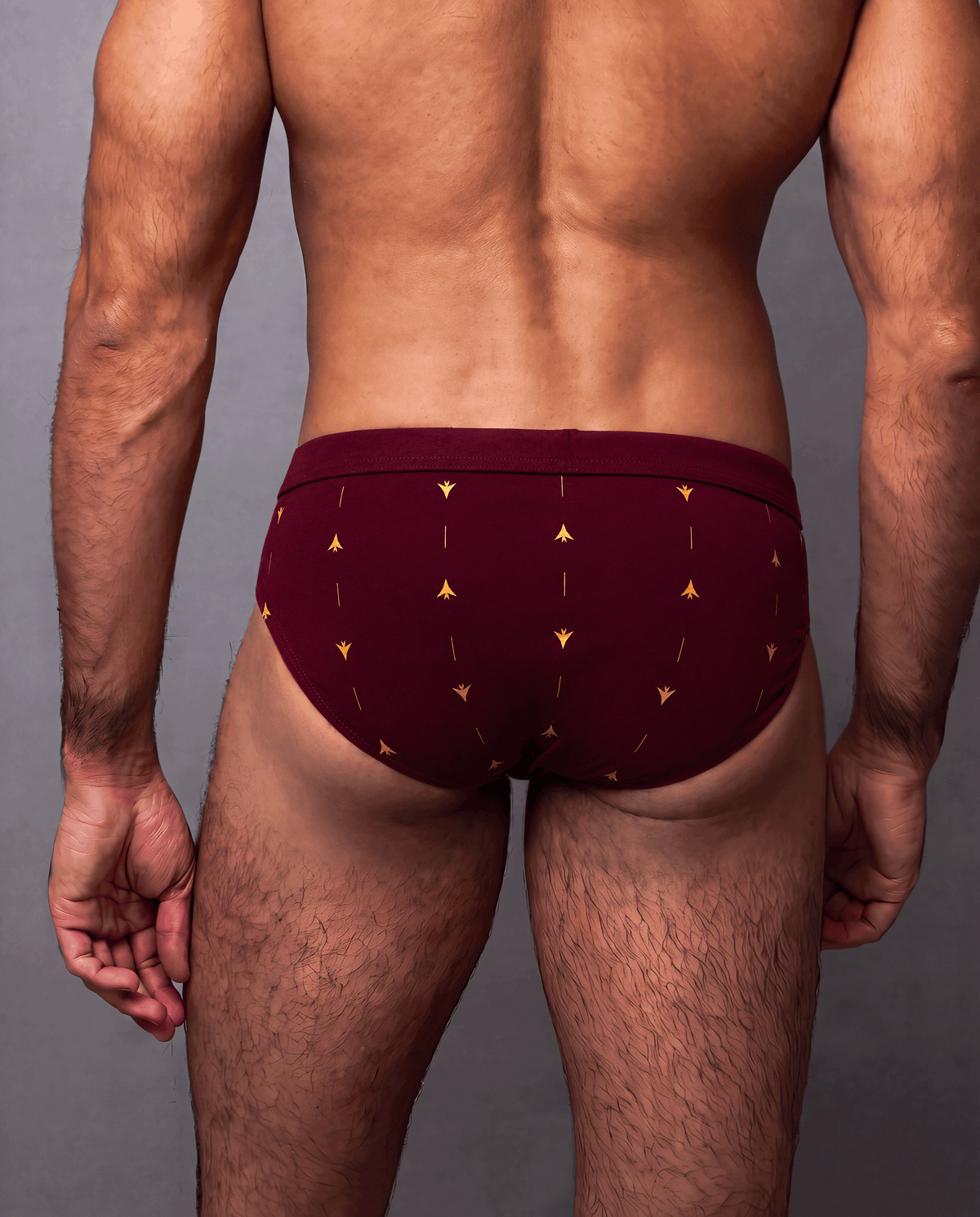 Rear view of a male model wearing the ELIO crimson low rise brief with gold print against a gray studio background.
