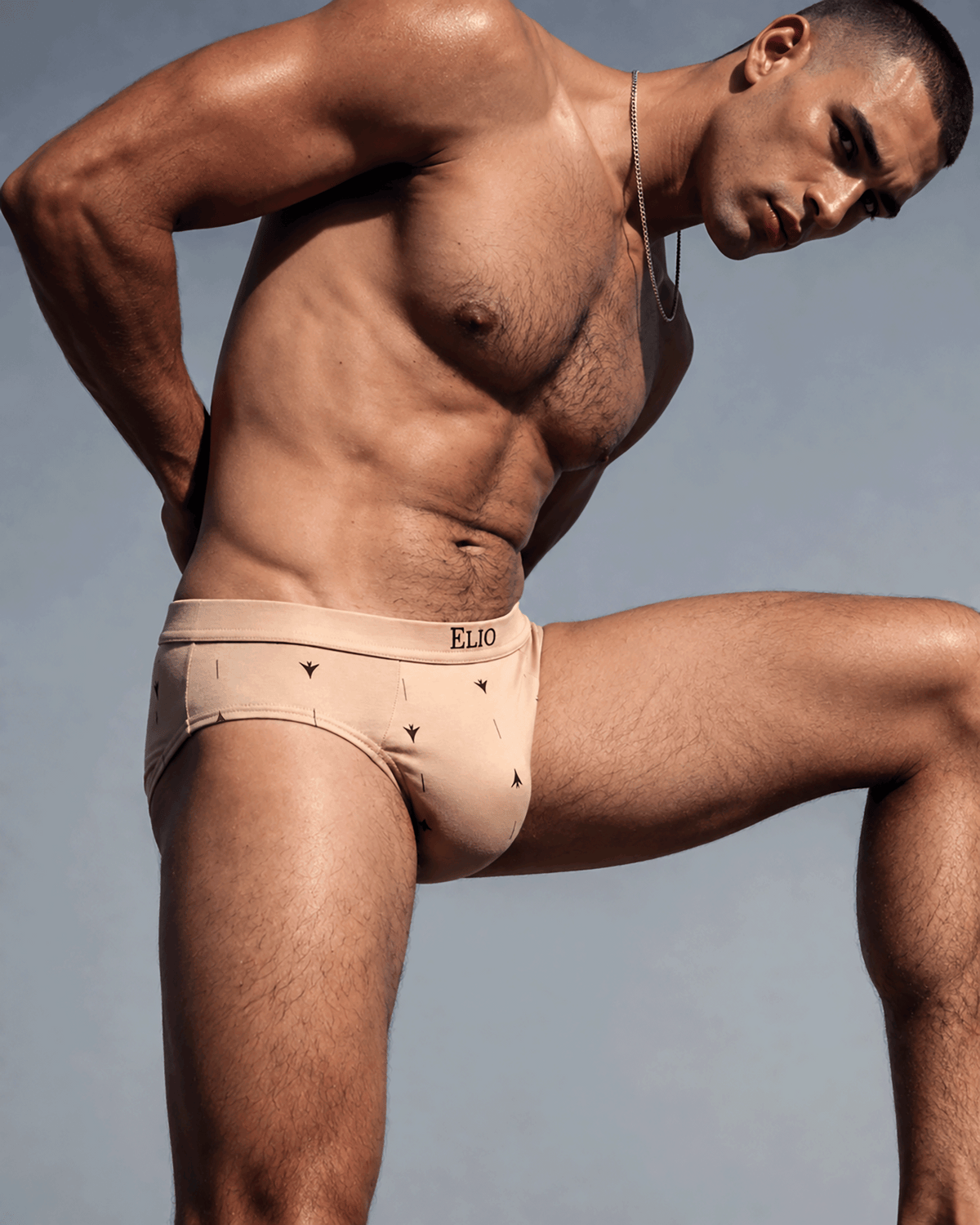 A muscular male model wearing beige ELJO branded briefs, posed dynamically against a solid light blue-gray background.